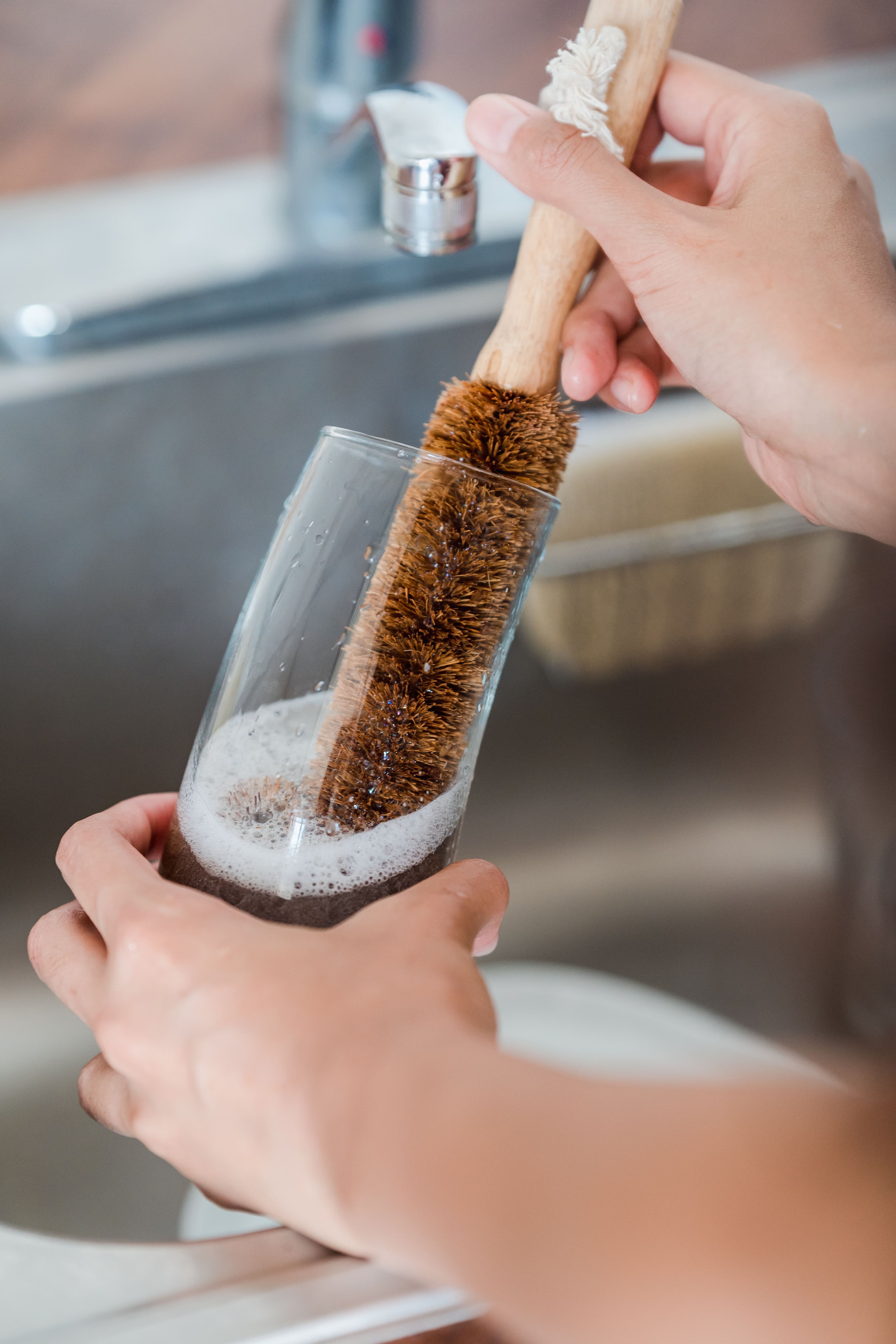 Hands using a long bottle brush inside a tall drinking glass over a sink